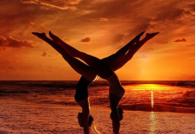 An image of two women balancing upside down on a beach in front of a sunset.