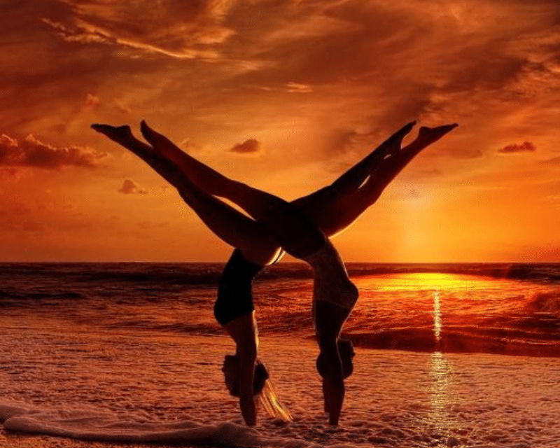 An image of two women balancing upside down on a beach in front of a sunset.