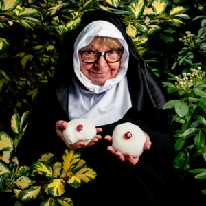 A promotional photo for Babs Horton’s play In the Lady Garden, showing Babs in a nun's habit holding two iced buns.